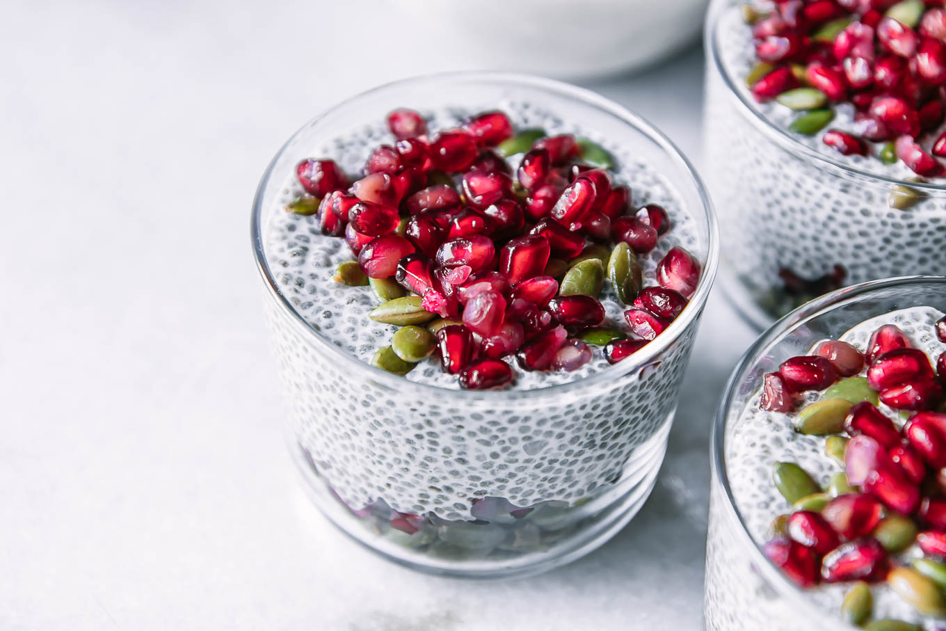 three small glass cups with pomegranate chia pudding and topped with seeds on a white table