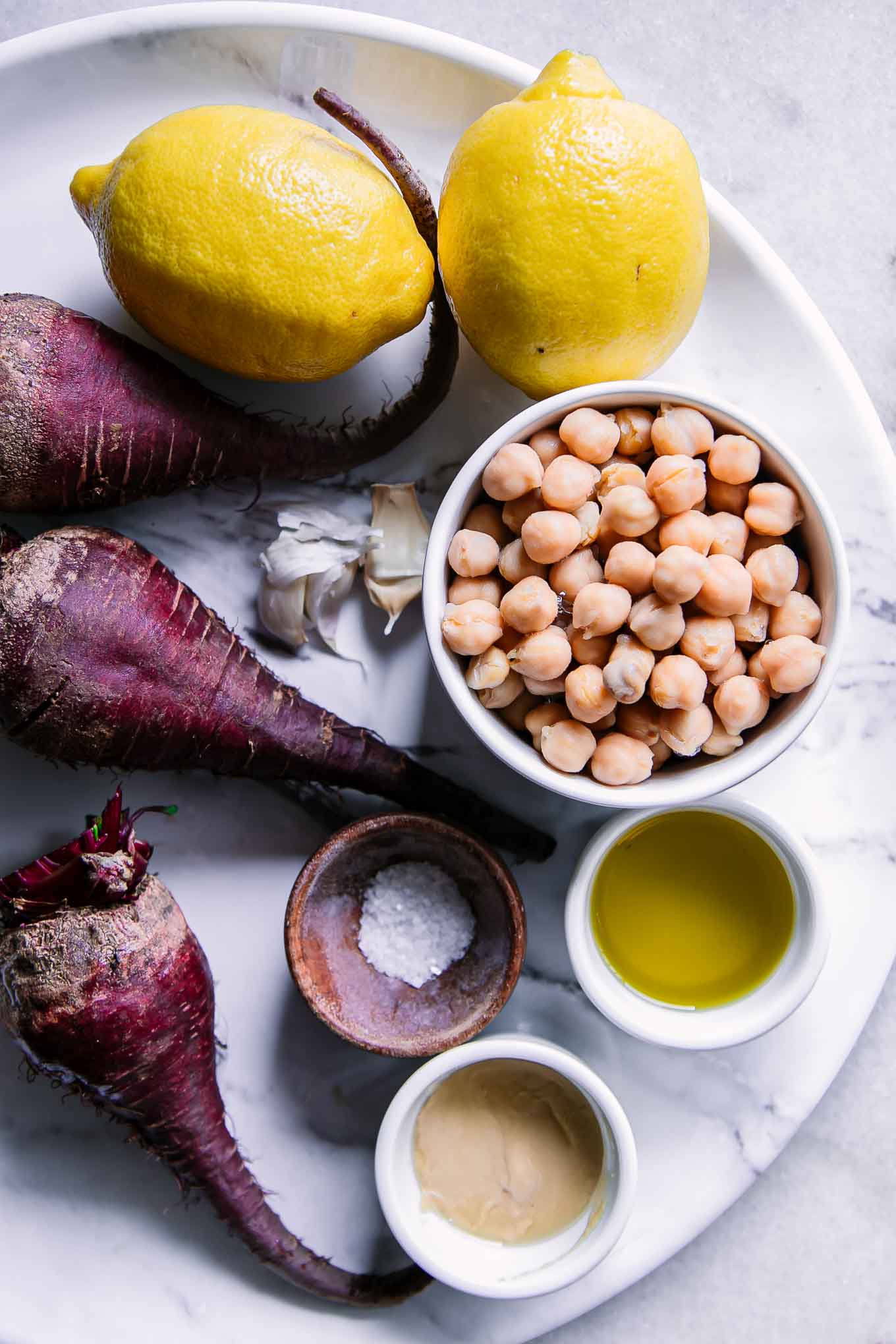 ingredients for red beet hummus including lemons, chickpeas, tahini, garlic, oil, salt, garlic, and beets on a white table