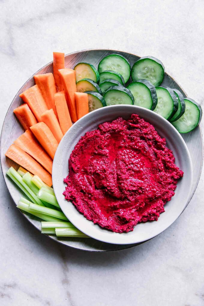 a bowl of red hummus with beets on a plate with cut cucumbers, carrots, and celery on a white table