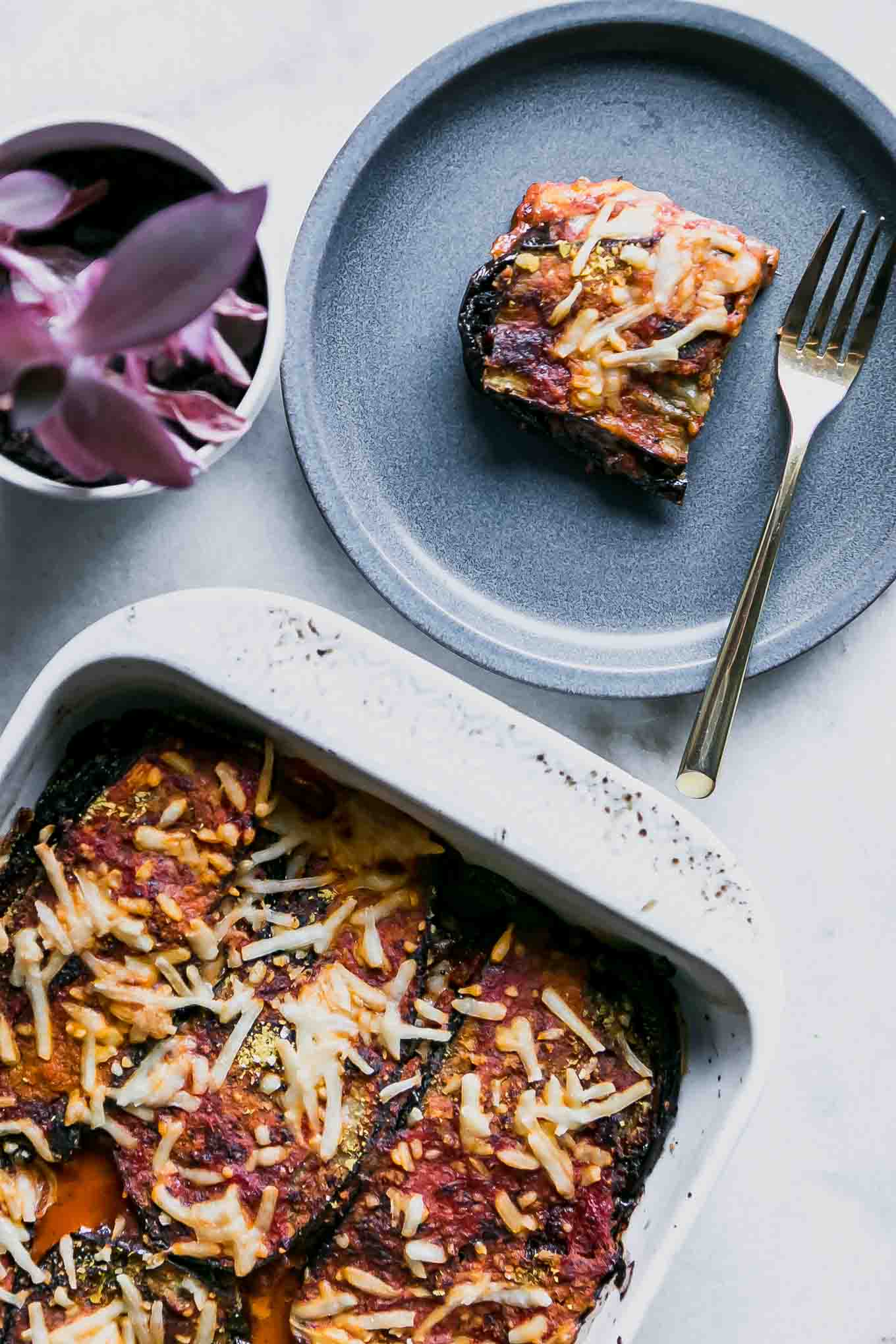 a cut piece of vegan eggplant parmesan on a blue plate with a baking dish full of baked eggplant parmesan on a white table