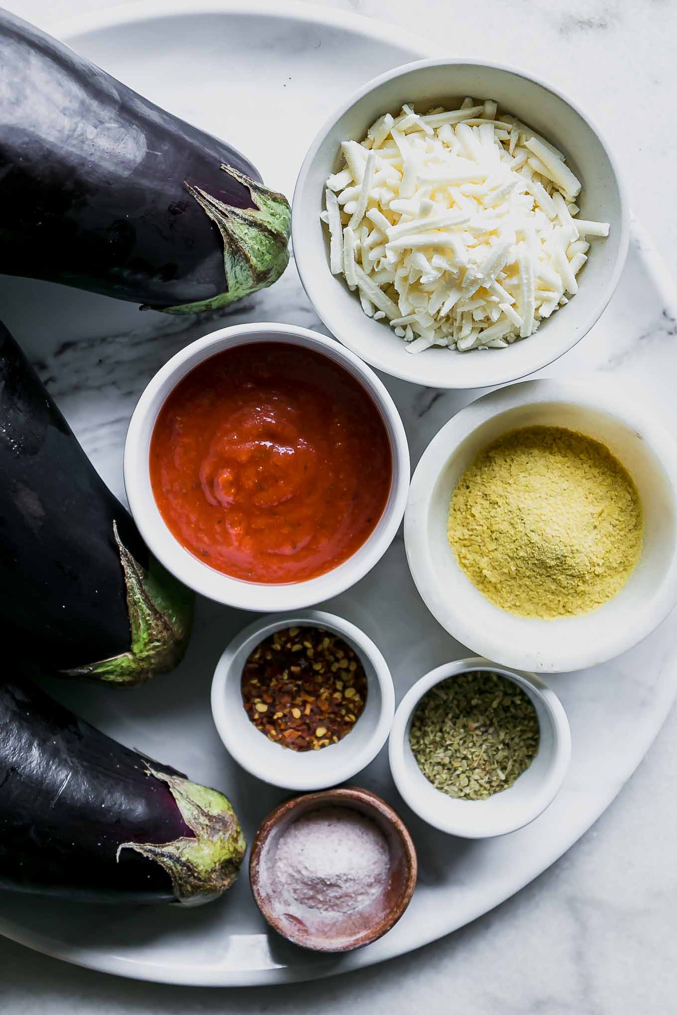 ingredients for eggplant parmesan on a white table, including eggplants, tomato sauce, and cheese