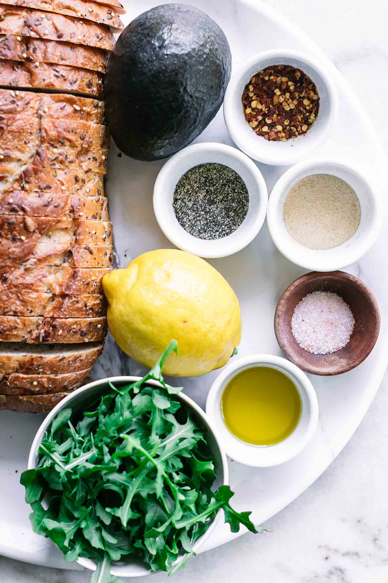 whole wheat bread, an avocado, a lemon, and bowls of arugula, oil, and seasonings on a white table for avocado toast
