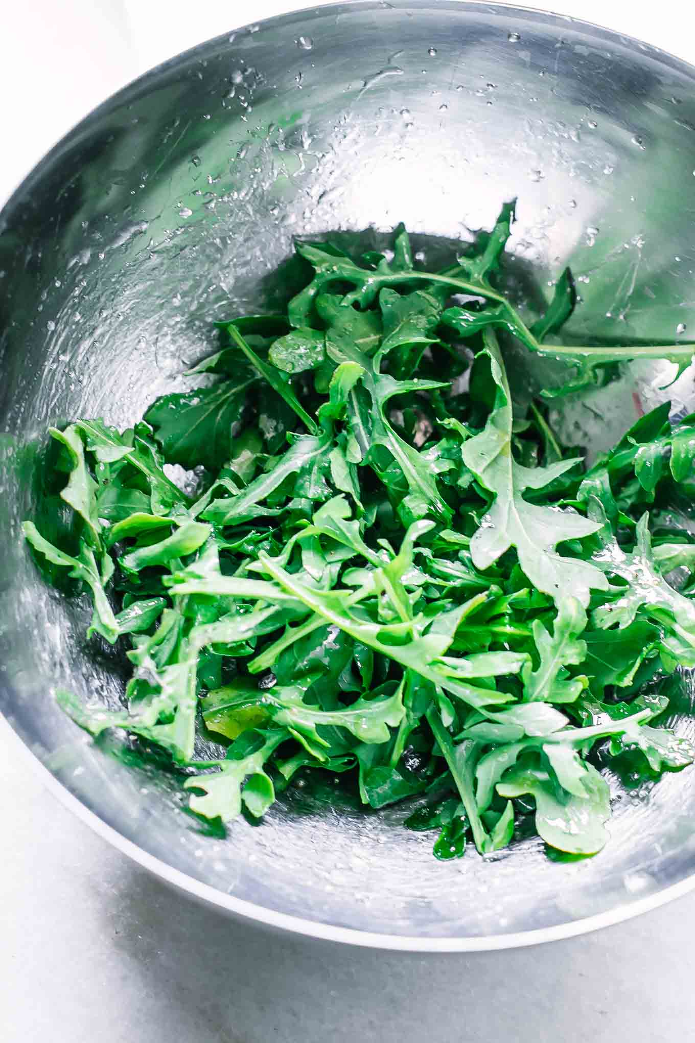 arugula with olive oil, lemon juice, and seasoning in a metal mixing bowl