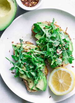 a white plate with avocado toast topped with fresh arugula with a half lemon