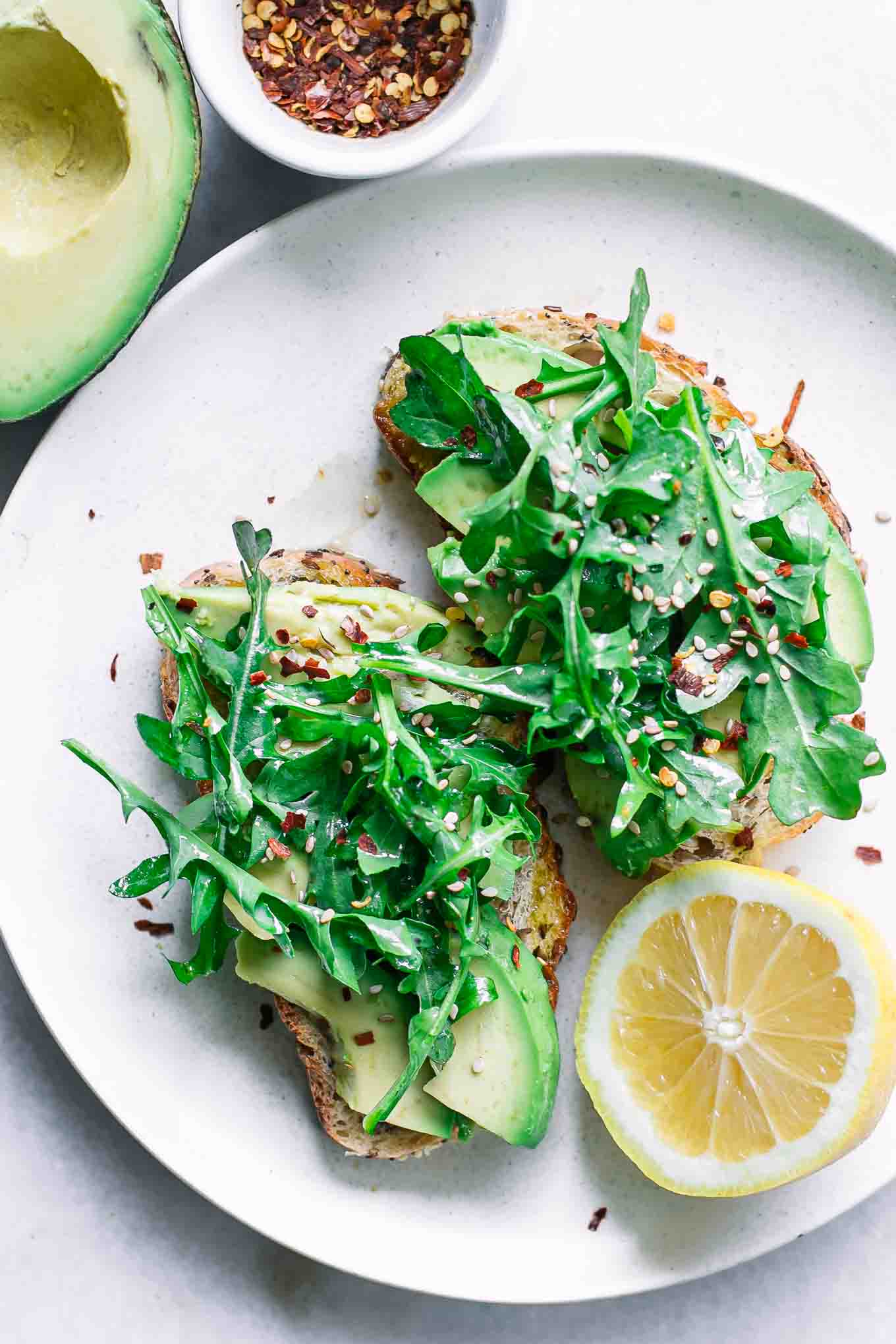 a white plate with avocado toast topped with fresh arugula with a half lemon