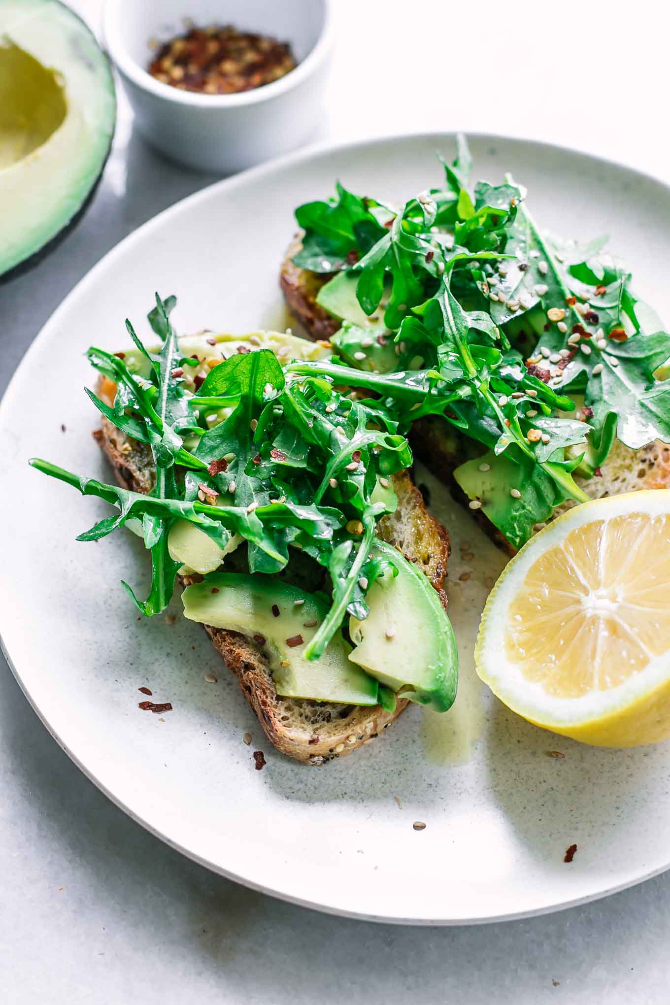 a white plate with arugula avocado toast and a lemon on a white table