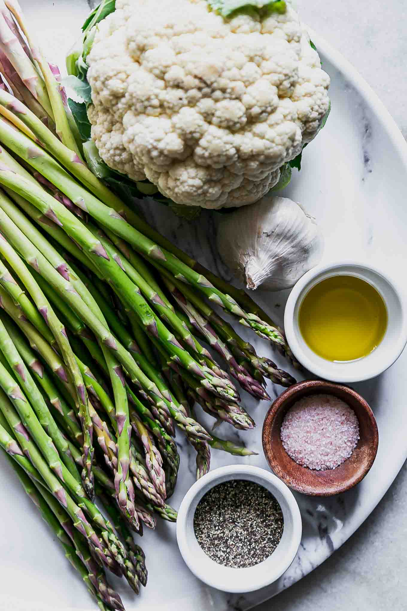 cauliflower, asparagus spears, garlic, and bowls of oil, salt, and pepper on a white table