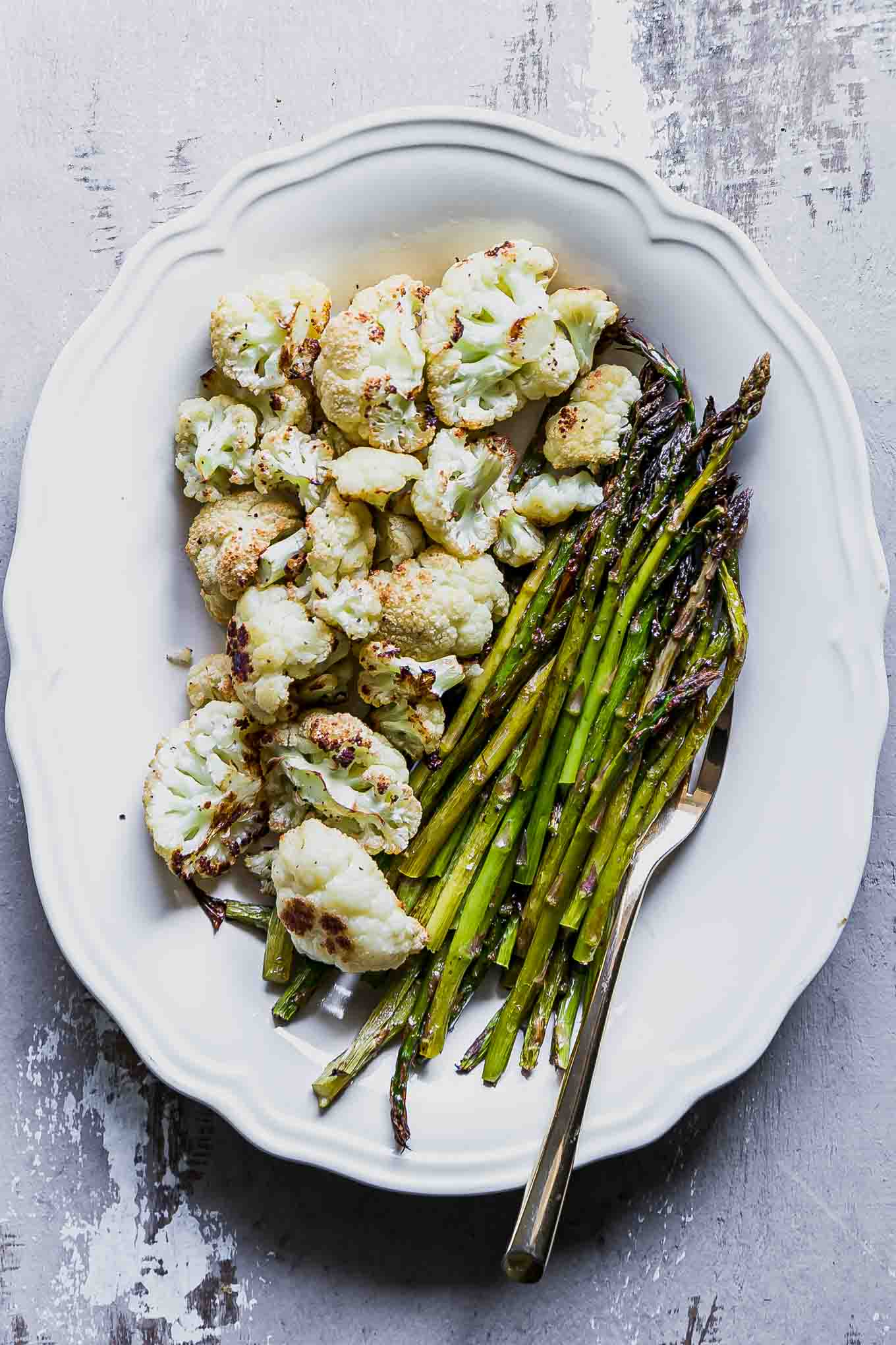 a white plate with baked asparagus and cauliflower and a gold serving fork on a wood table