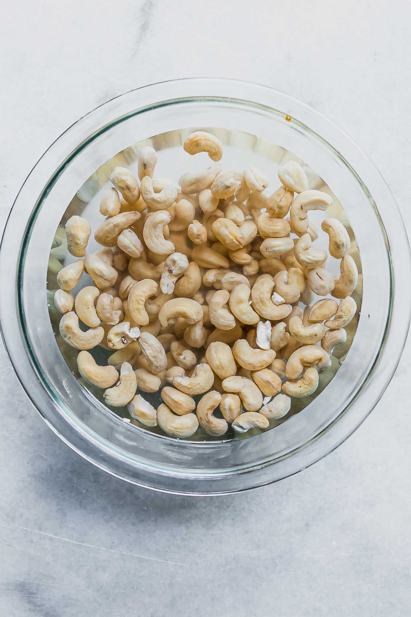 a glass mixing bowl with cashews soaking in hot water on a white table