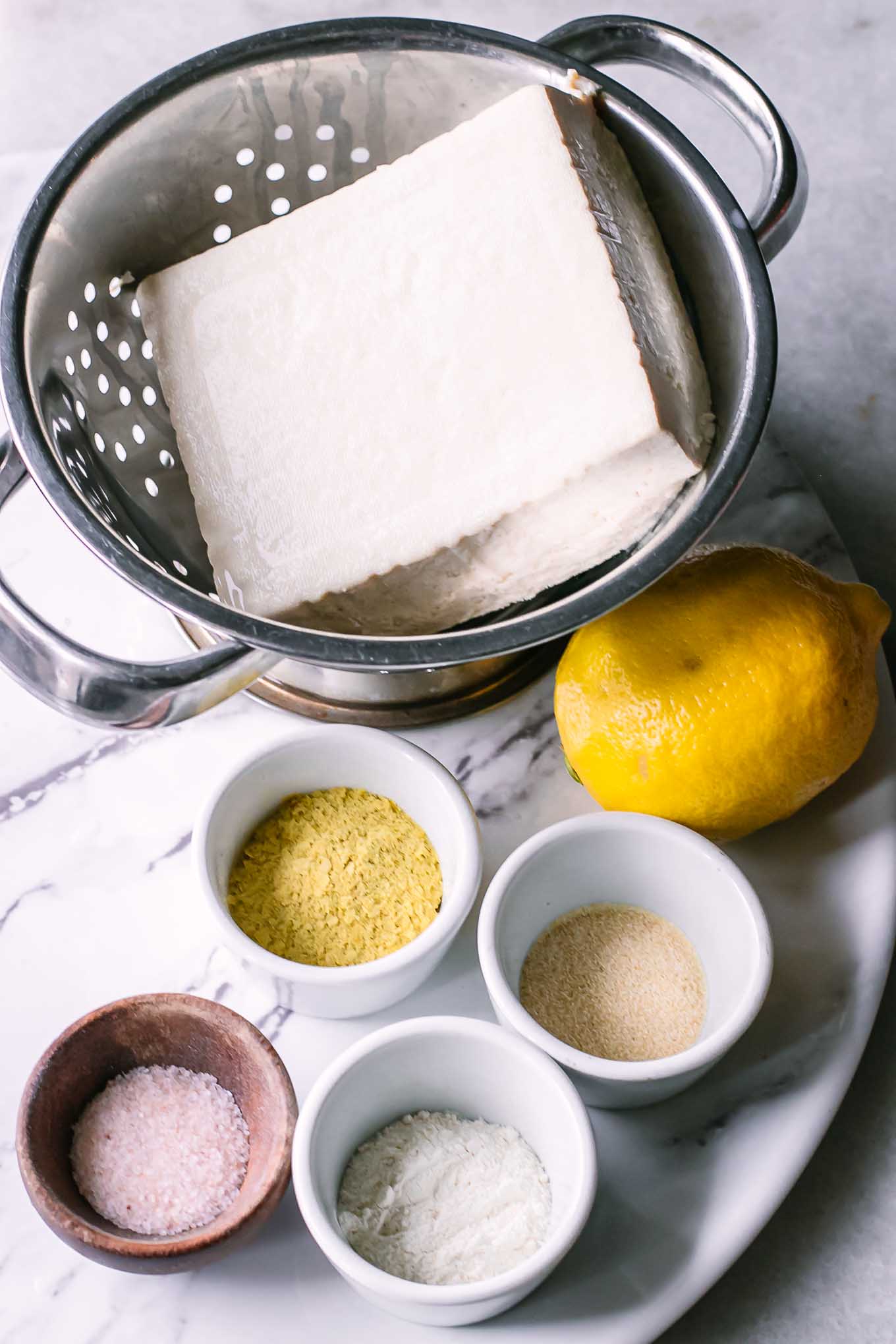 a colander with a block of tofu, a lemon, and bowls of salt, nutritional yeast, garlic powder, and onion powder for vegan ricotta cheese