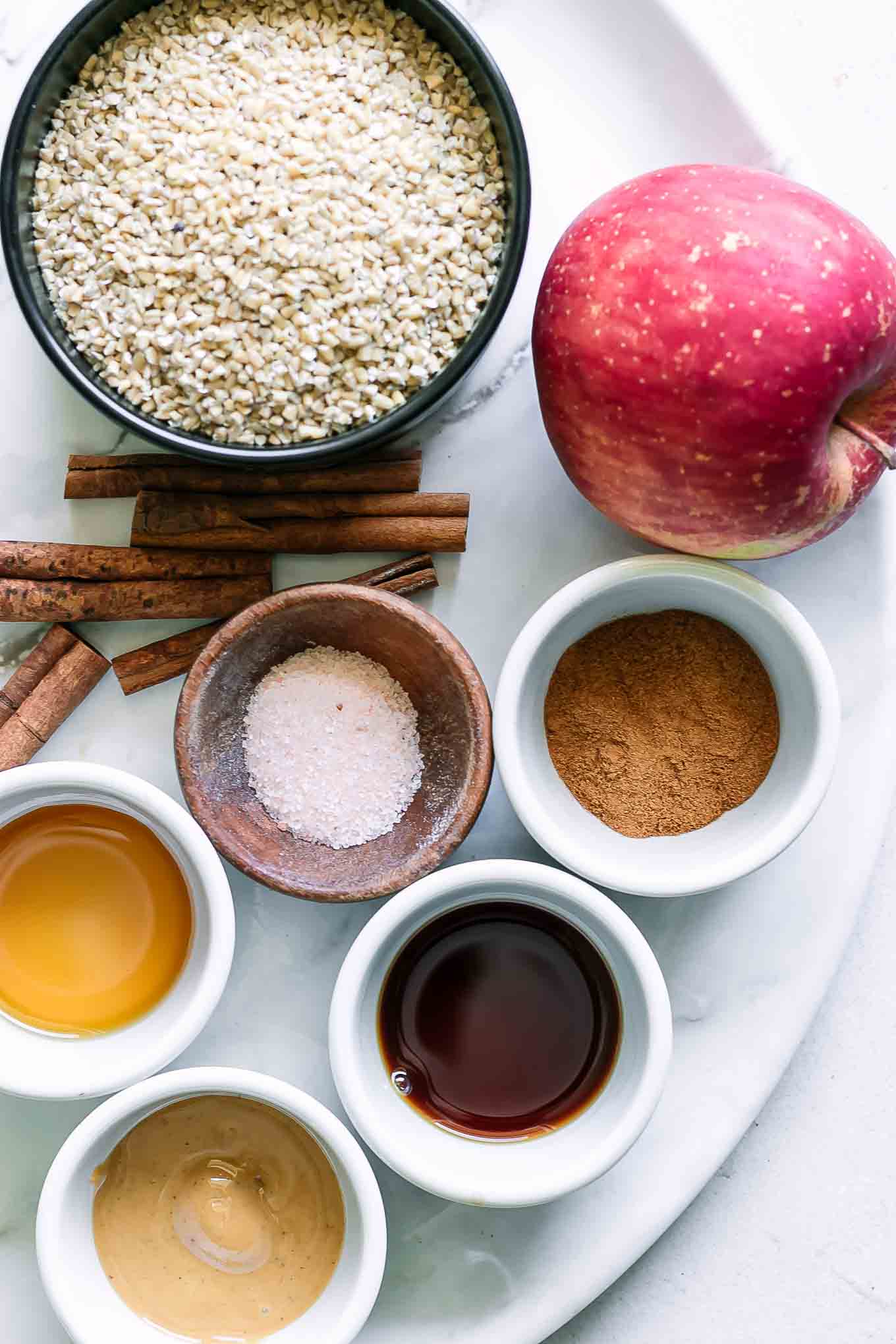 bowls of oatmeal, cinnamon, vanilla, maple syrup, spices, and an apple on a table