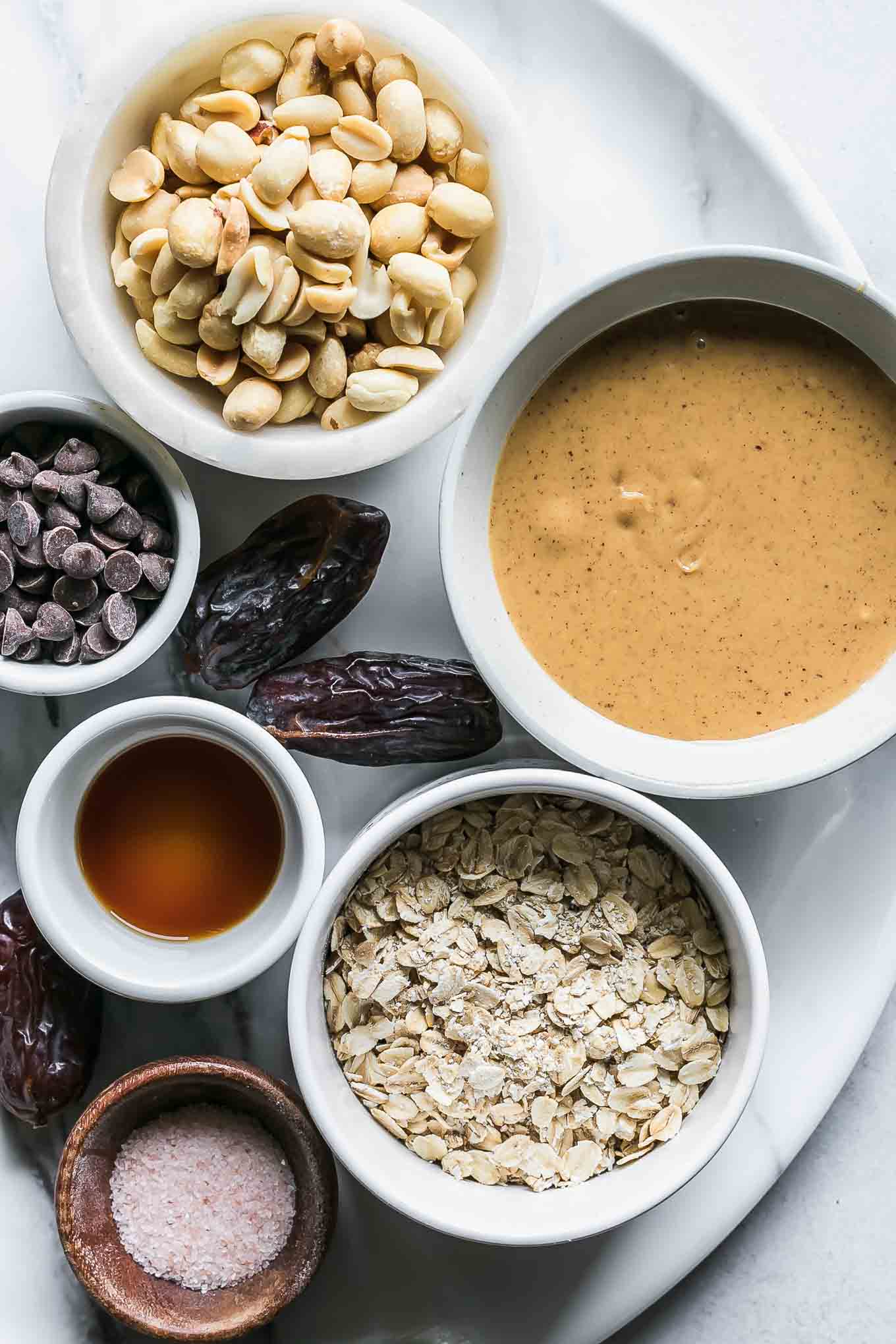 bowls of peanuts, peanut butter, rolled oats, chocolate chips, vanilla, and salt for granola bars on a white table