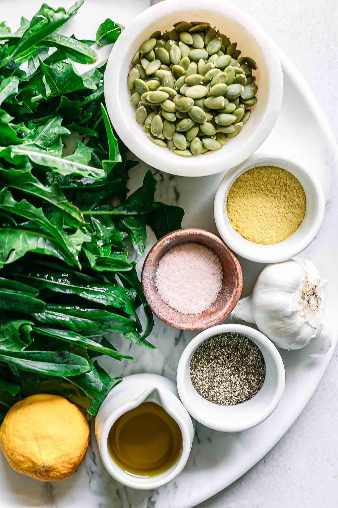 fresh dandelion leaves on a table with bowls of nuts, oil, cheese, and seasonings for pesto sauce