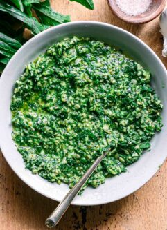 dandelion greens pesto in a white bowl with a gold fork on a wood table with fresh dandelion leaves