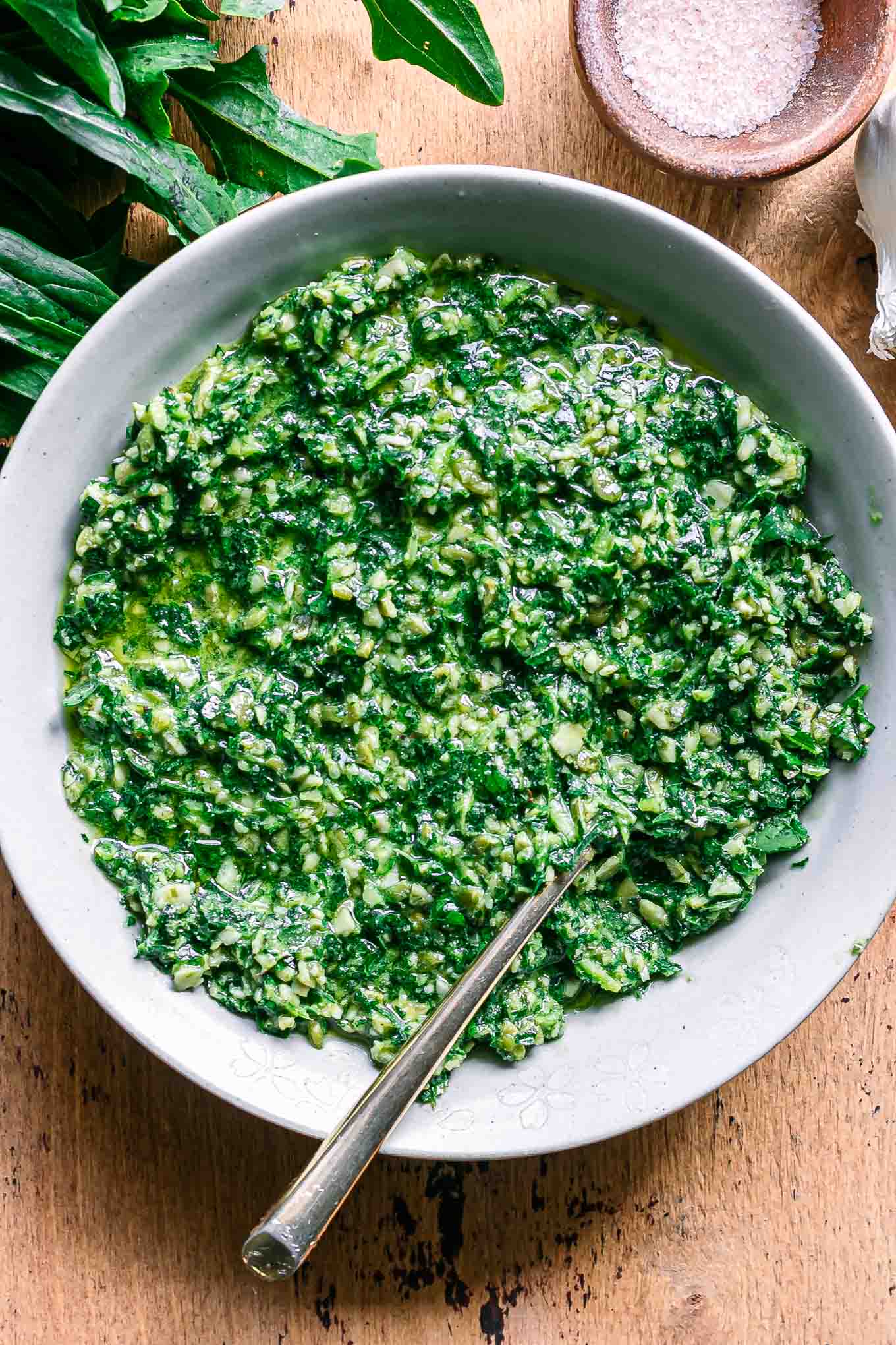 dandelion greens pesto in a white bowl with a gold fork on a wood table with fresh dandelion leaves