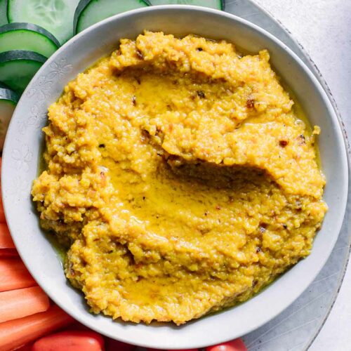 a bowl of orange-colored golden beet hummus dip with cut vegetables for dipping on a white table