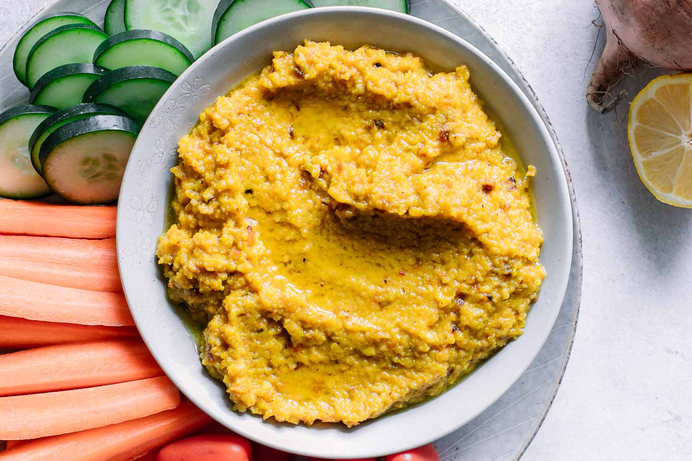 a bowl of orange-colored golden beet hummus dip with cut vegetables for dipping on a white table