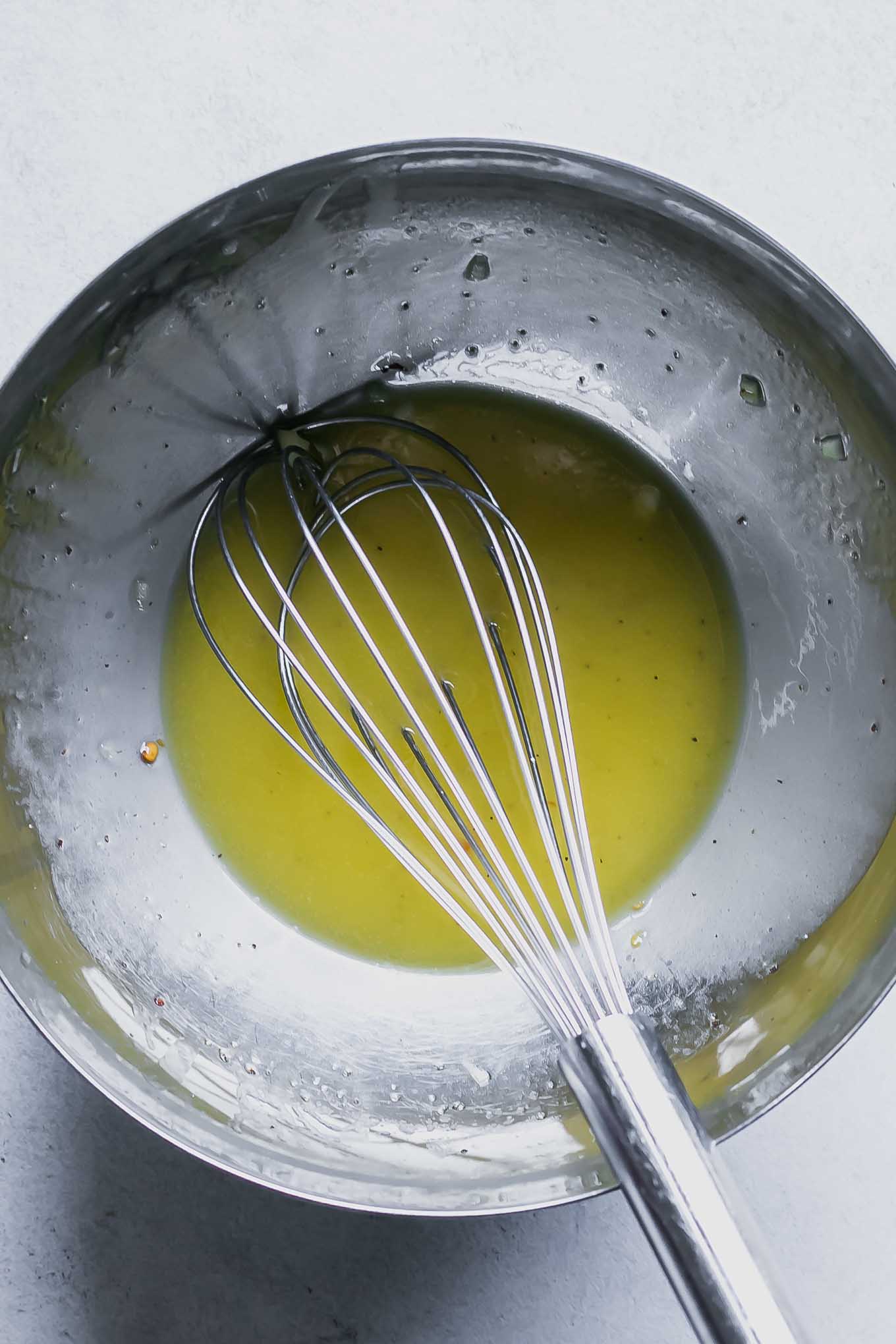 a bowl of olive oil with lemon juice and minced garlic with a whisk on a white table