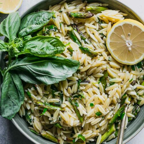 a bowl of lemon asparagus orzo with fresh herbs on a white table with a lemon