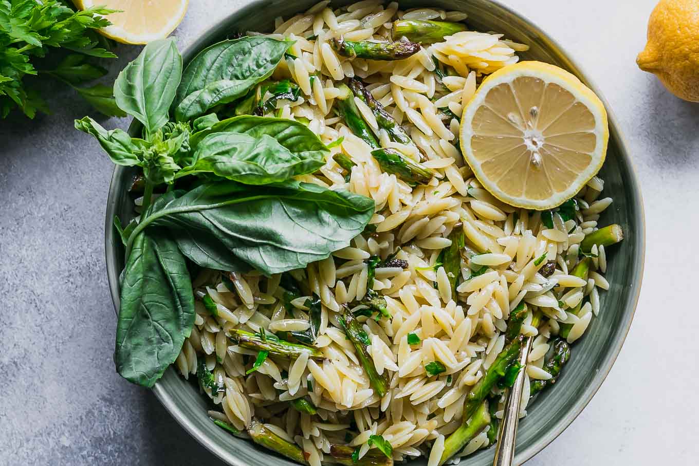 a bowl of lemon asparagus orzo with fresh herbs on a white table with a lemon