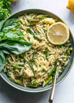 a bowl of orzo pasta with asparagus and fresh herbs with a lemon on a table with a gold fork