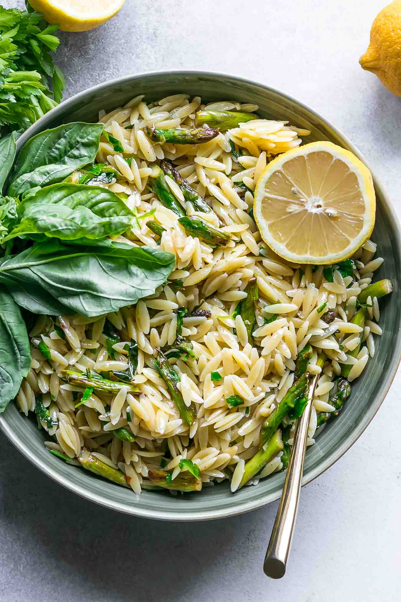 a bowl of orzo pasta with asparagus and fresh herbs with a lemon on a table with a gold fork