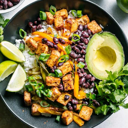 a bowl with a tofu fajita bowl with avocado, limes, and cilantro on a white table