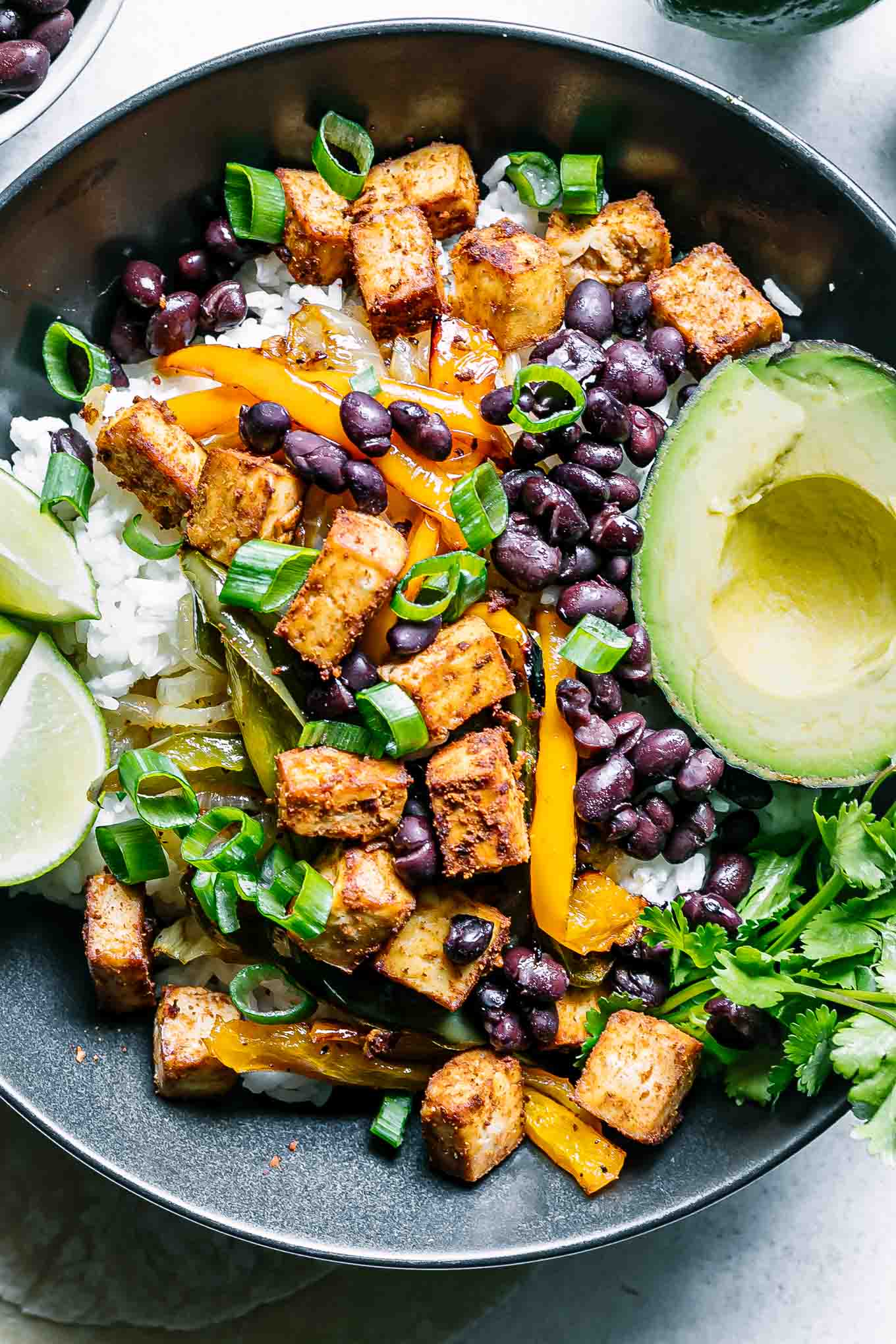 a close up photo of a black bowl with tofu, black beans, roasted fajita vegetables, limes, and avocado on a white table