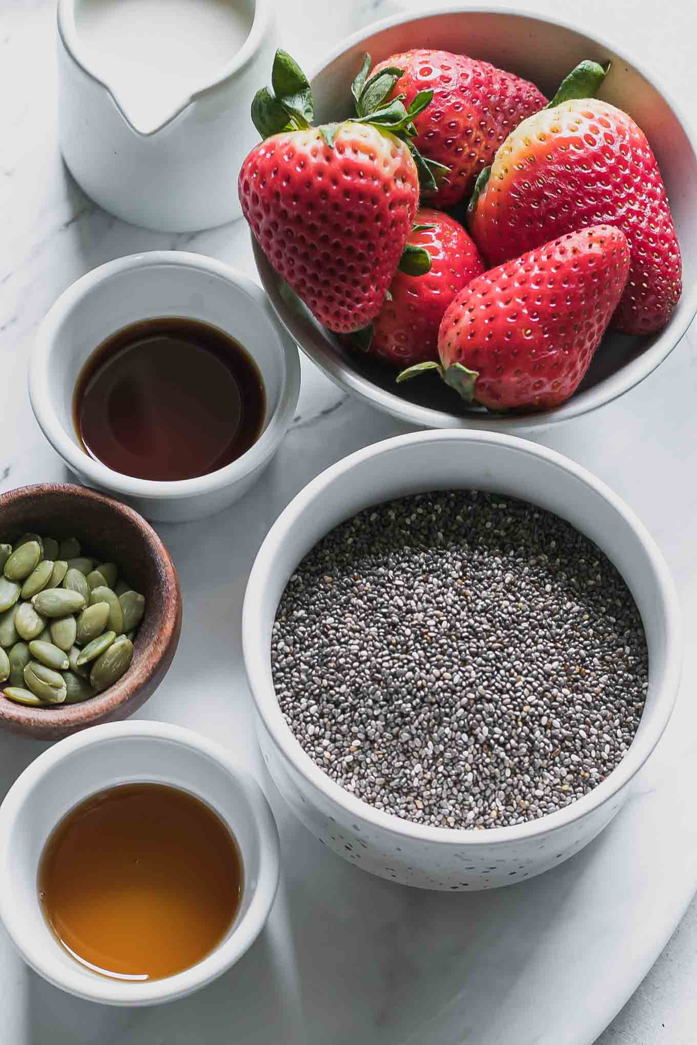 bowls of chia seeds, strawberries, vanilla, maple syrup, plant-based milk, and seeds for chia pudding on a white table