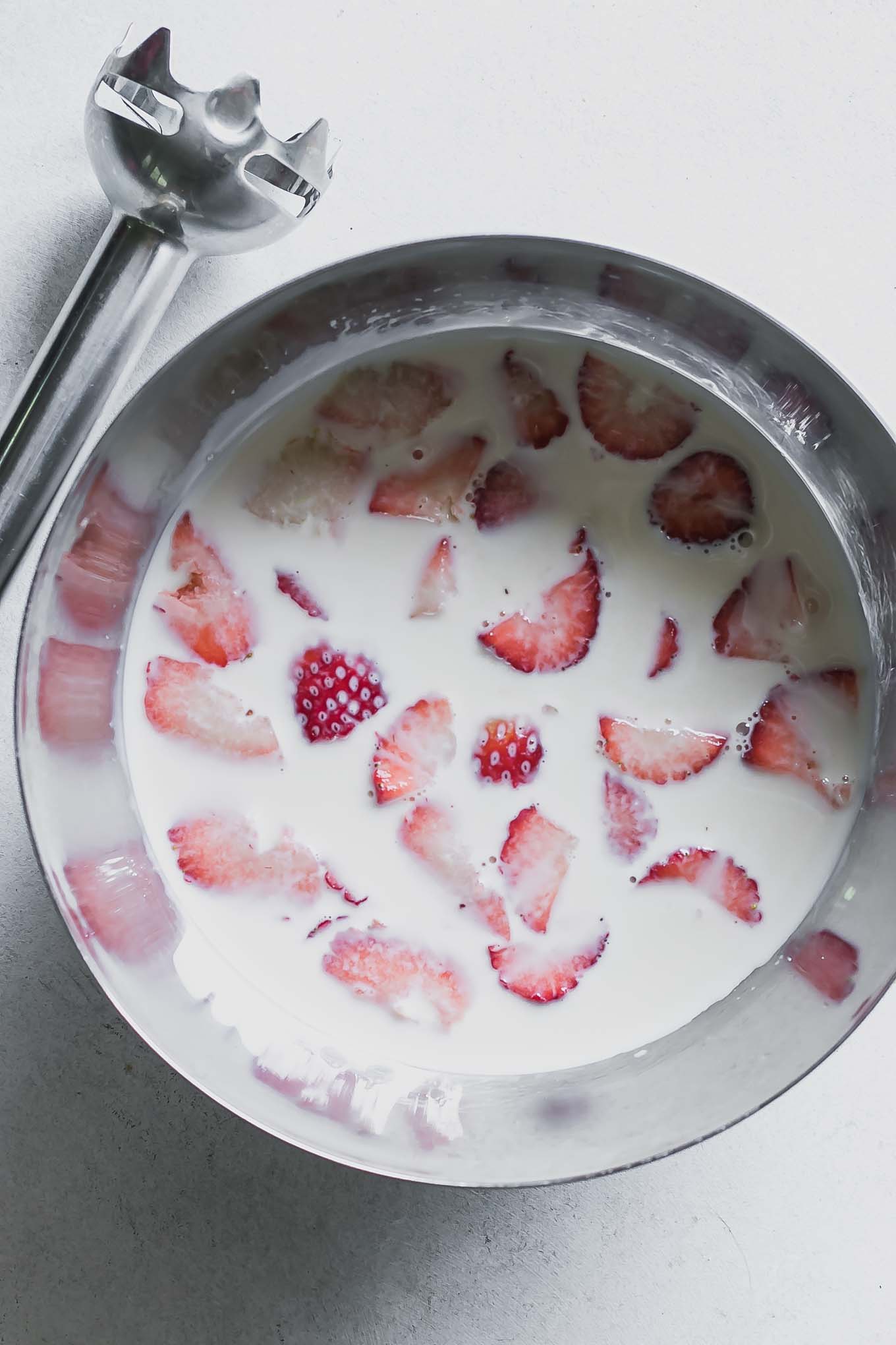 a metal mixing bowl with milk and sliced strawberries and a handheld immersion blender on a white table