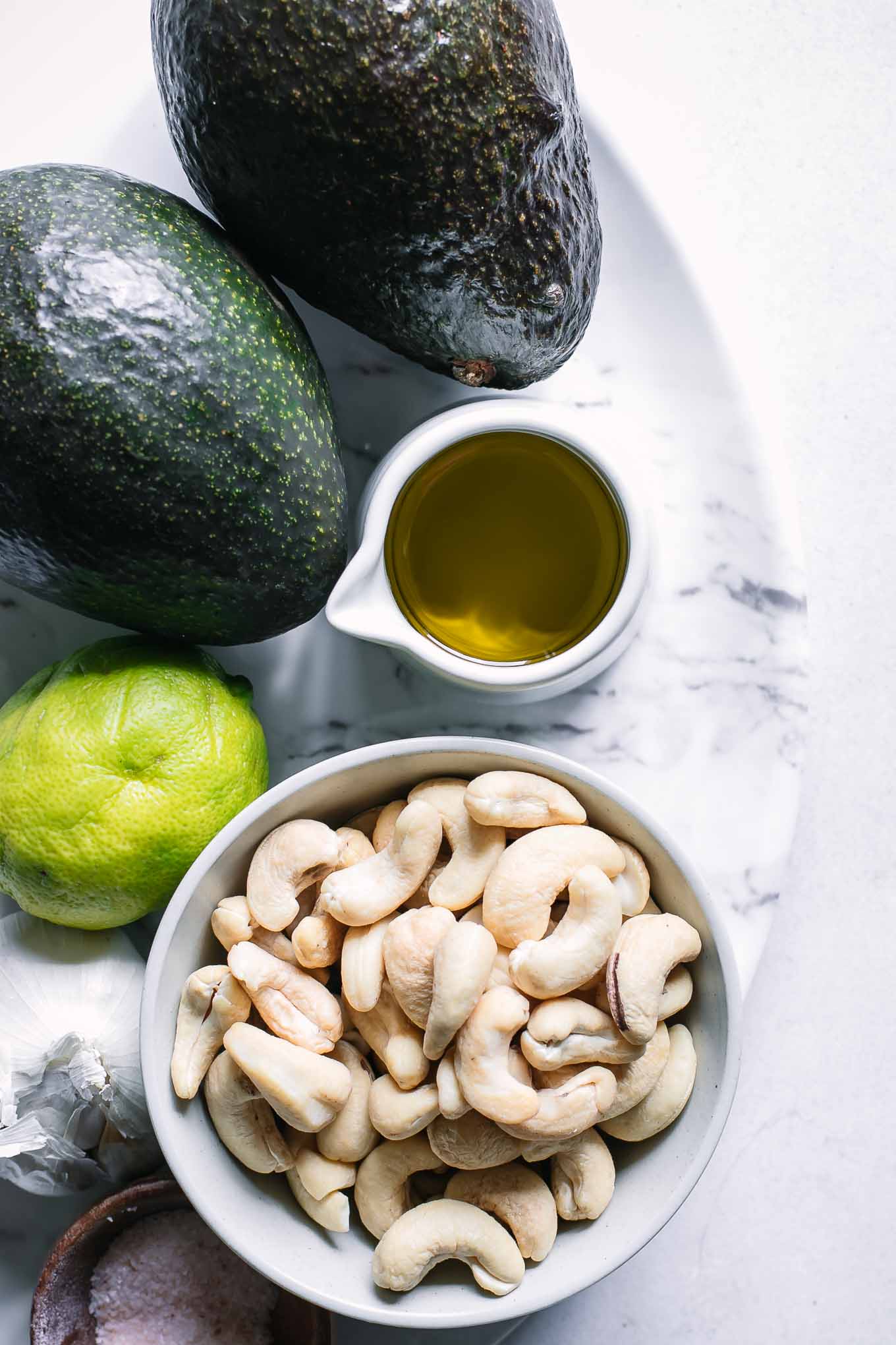 avocados, lime juice, cashews, oil, garlic, and salt on a white table for avocado sauce