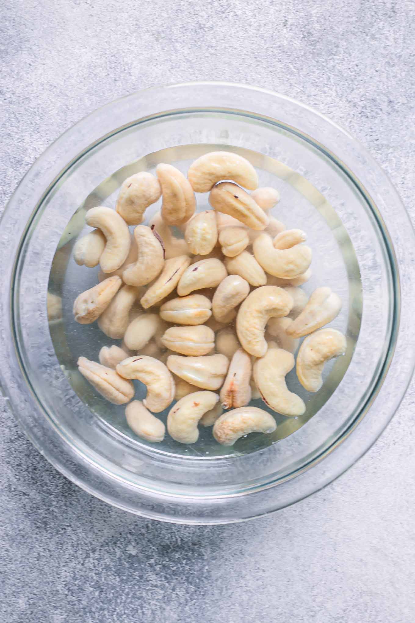 a glass mixing bowl with cashews soaking in hot water on a blue table