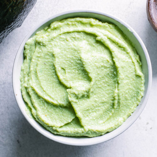 a bowl of vegan avocado crema sauce on a white table with an avocado