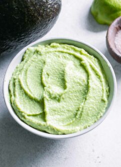 a bowl of vegan avocado crema sauce in a white bowl with an avocado and lime