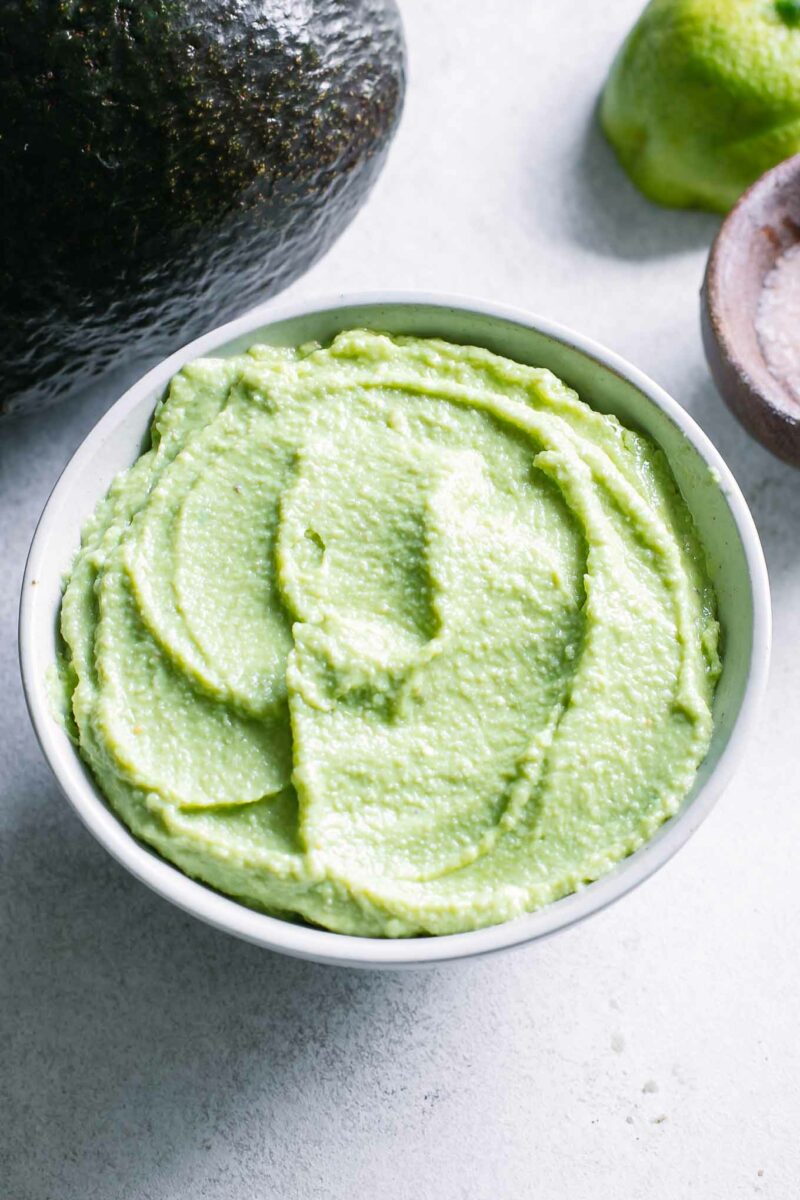 a bowl of vegan avocado crema sauce in a white bowl with an avocado and lime