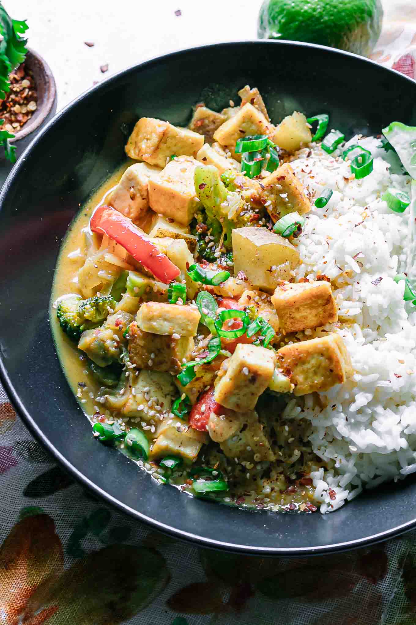 a close up photo of yellow curry with tofu and potatoes with white rice in a black bowl