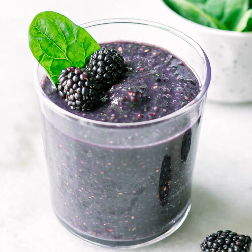 a spinach blackberry smoothie in a glass on a white table with fresh blackberries