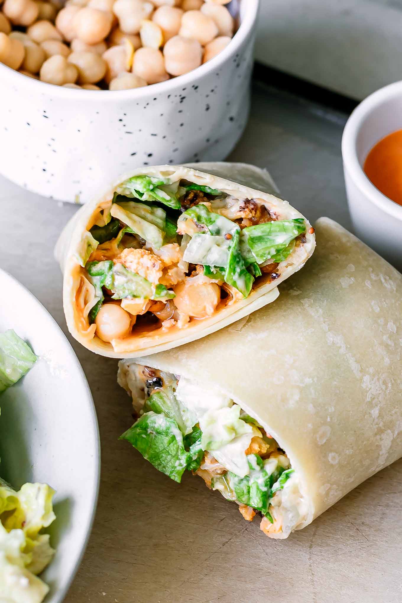 two buffalo caesar wrap halves on a table with a bowl of chickpeas and a small bowl of buffalo sauce