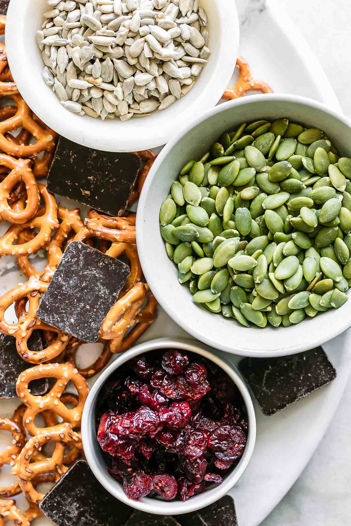 bowls of pretzels, dark chocolate, dried cranberries, pumpkin seeds, and sunflower seeds on a white table for trail mix