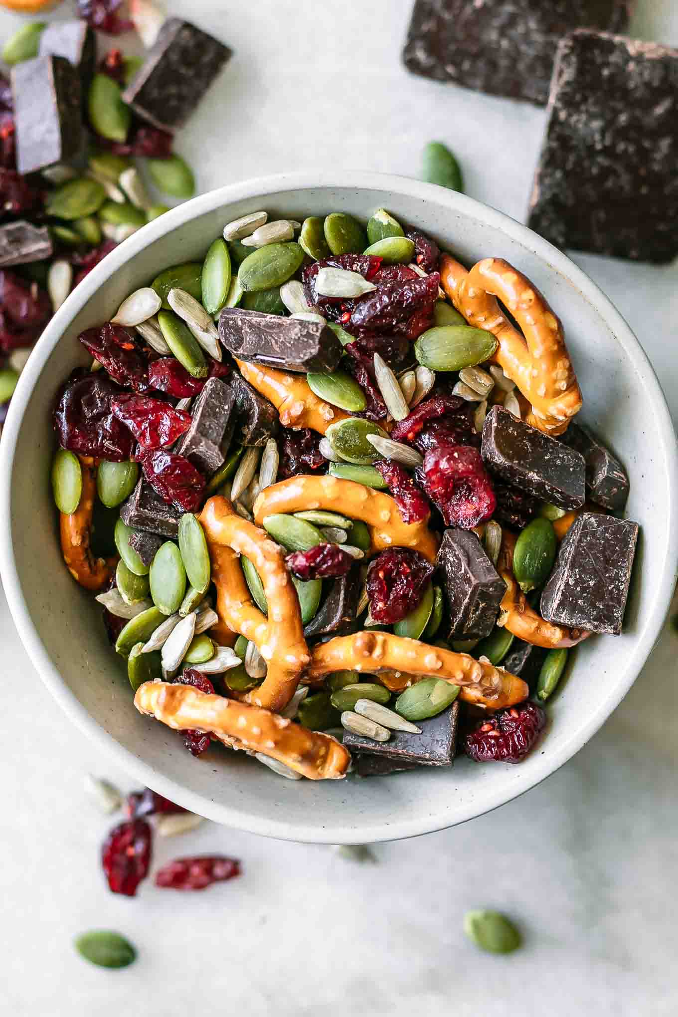 a close up photo of a small bowl with trail mix with dark chocolate, dried cranberries, pretzels, pumpkin seeds, and sunflower seeds