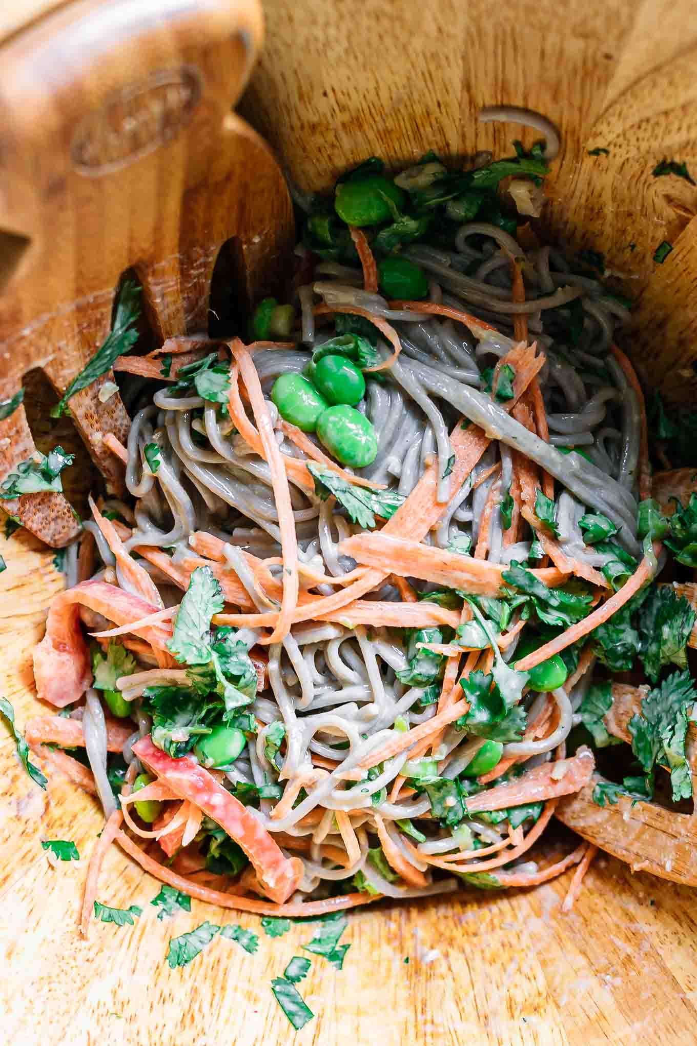 a close up of a bowl of soba noodle salad with wood salad tongs