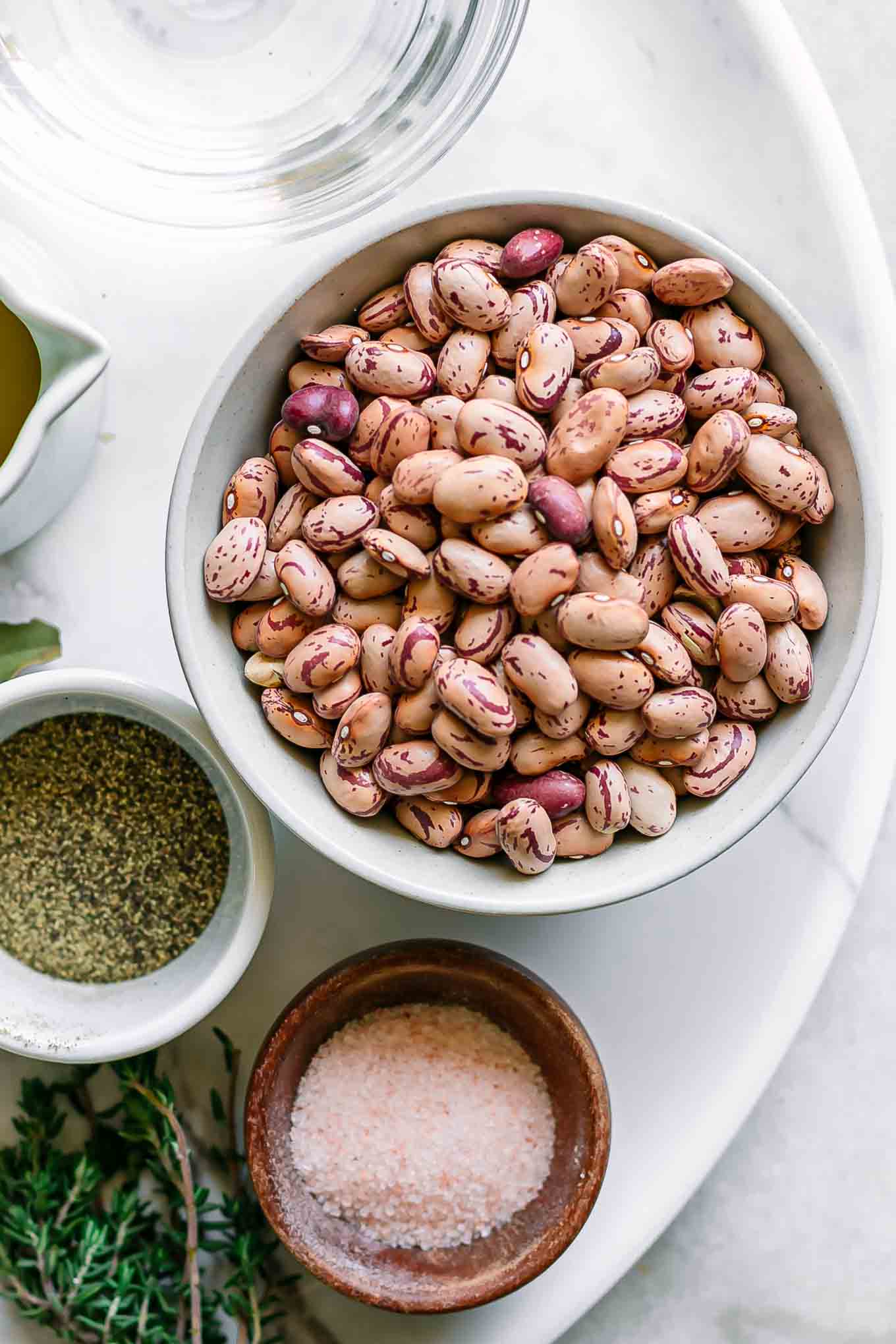 a bowl of dried cranberry beans on a table with salt, pepper, water, oil, and herbs