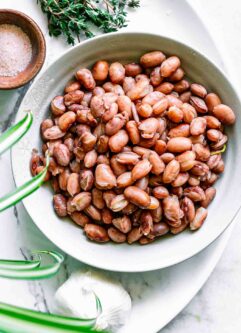 a bowl of cooked cranberry beans on a white table with herbs and spices