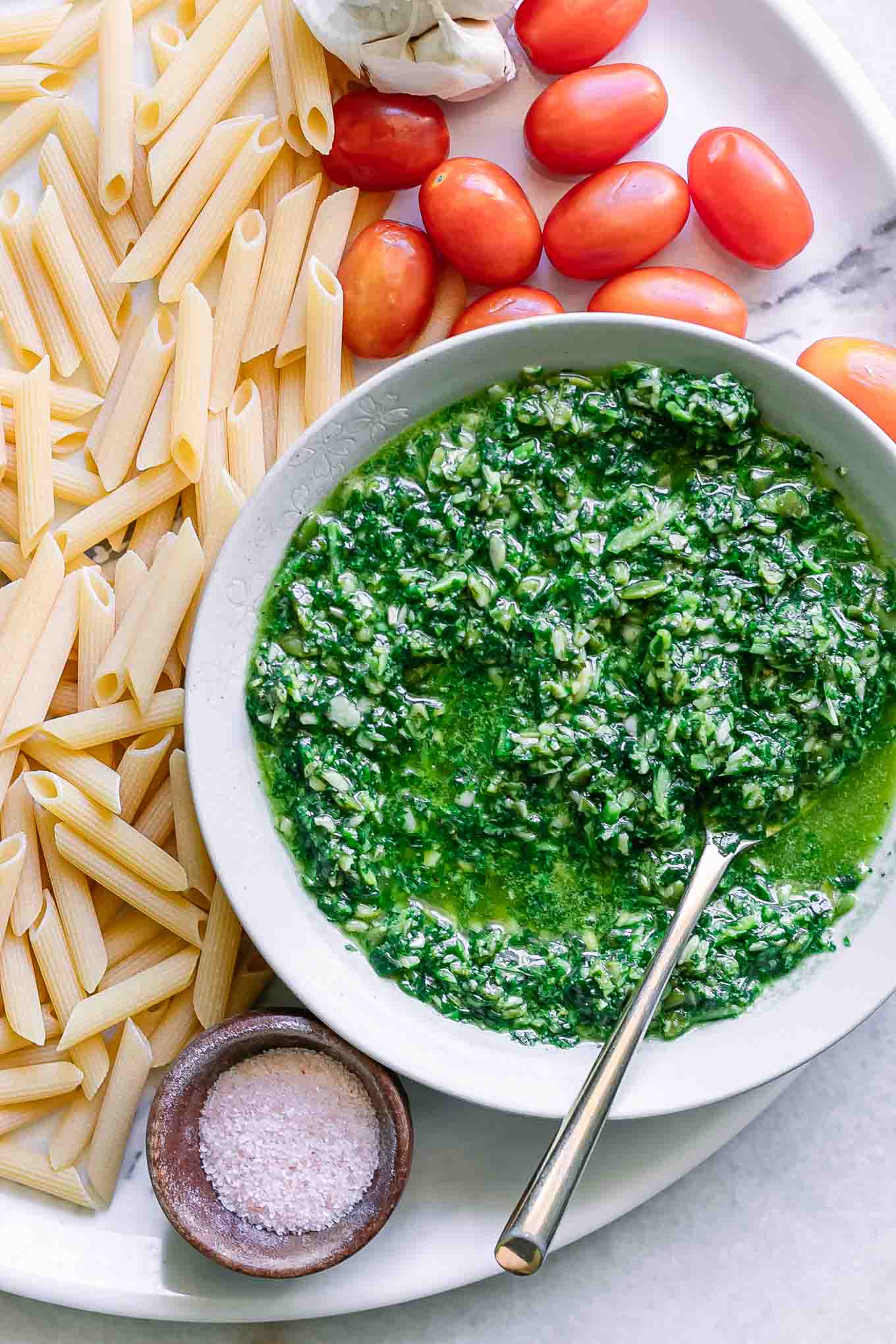 a bowl of pesto sauce on a table with cherry tomatoes, dry penne pasta, garlic, and salt