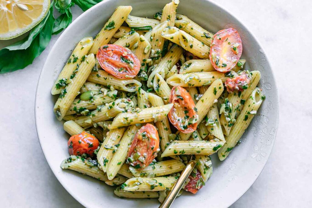 a white bowl with pesto penne pasta and cherry tomatoes on a white table with a gold fork