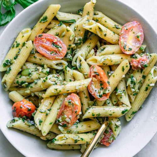 a white bowl with pesto penne pasta and cherry tomatoes on a white table with a gold fork