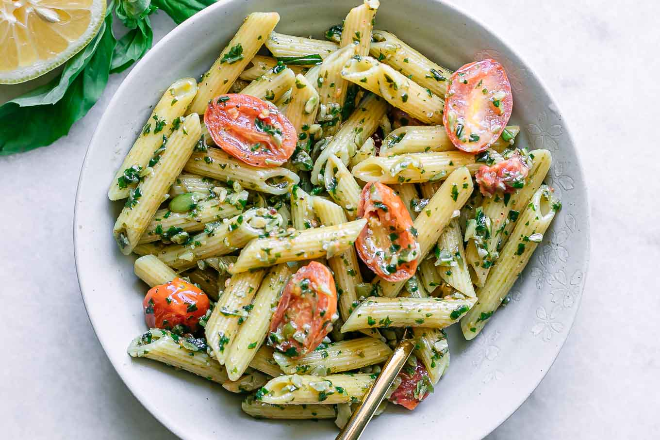 a white bowl with pesto penne pasta and cherry tomatoes on a white table with a gold fork