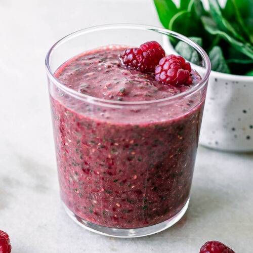 a glass with a spinach raspberry smoothie on a white table with fresh strawberries and a bowl of fresh spinach