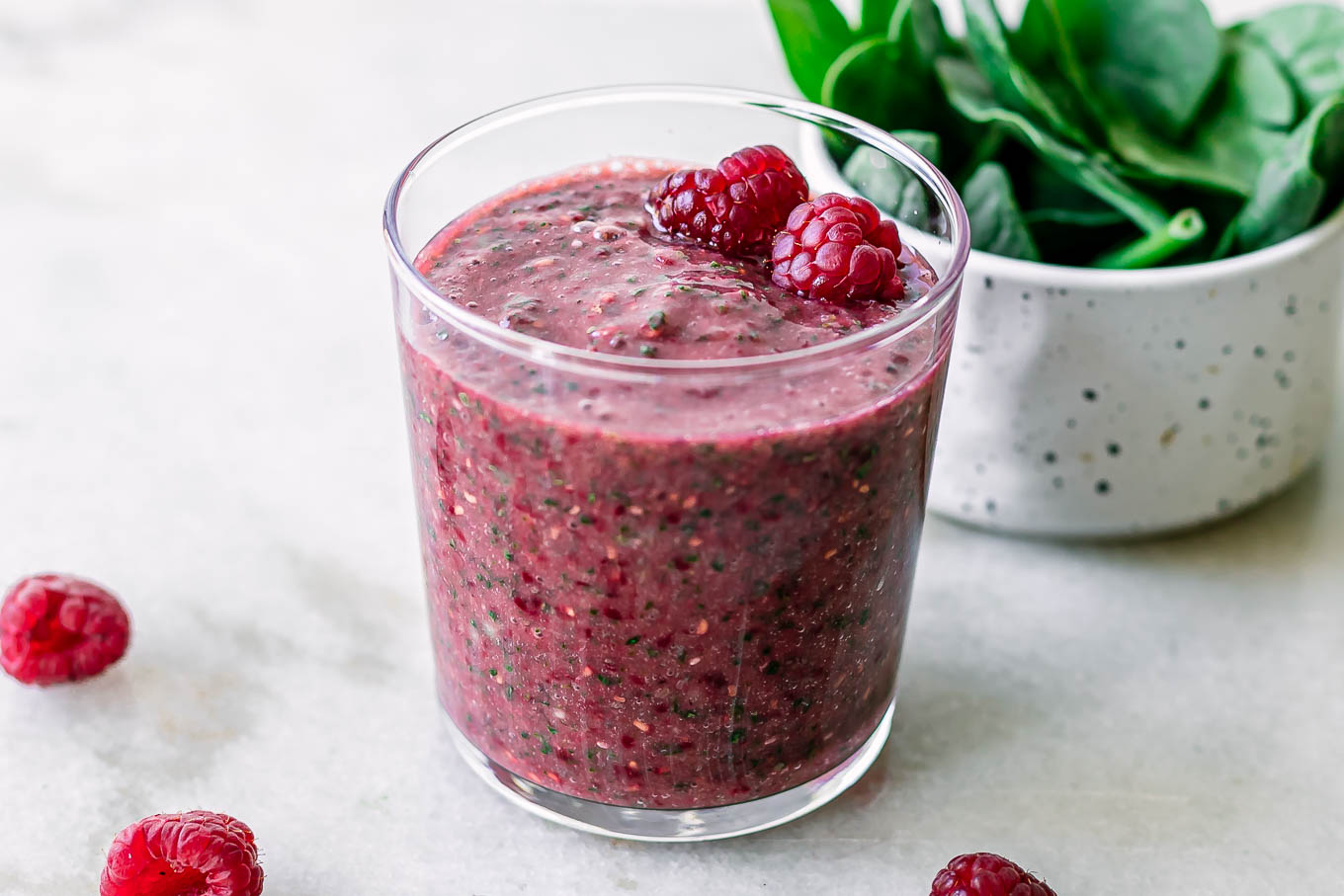 a glass with a spinach raspberry smoothie on a white table with fresh strawberries and a bowl of fresh spinach