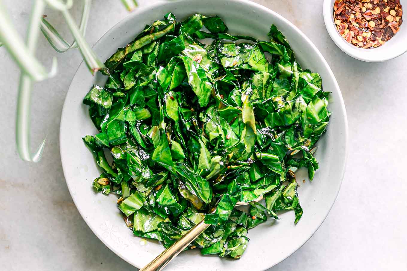 a bowl of sautéed collard greens on a white table with bowls of spices